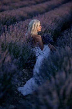 Blonde woman poses in lavender field at sunset. Happy woman in white dress holds lavender bouquet. Aromatherapy concept, lavender oil, photo session in lavender.