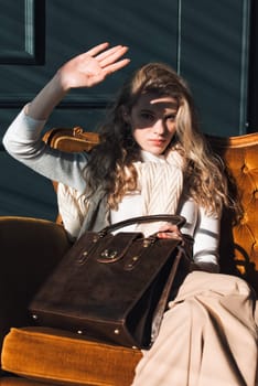 beautiful curly blond hair woman posing with a small shopper brown bag in a vintage chair. Model wearing stylish white sweater and classic trousers