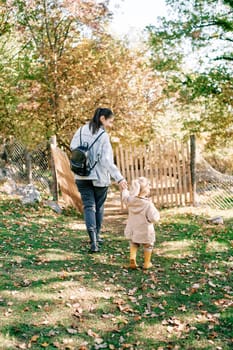 Mother and a little girl are walking holding hands along a path in the park to a wooden gate. Back view. High quality photo