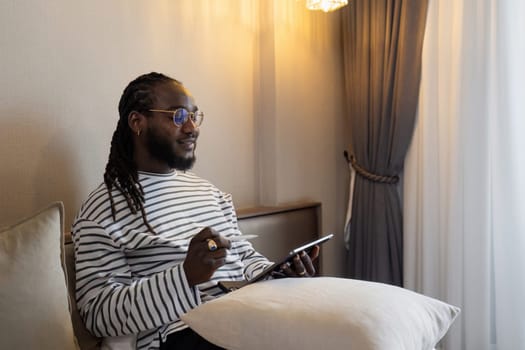 African American business man working at home on weekend, holding tablet while working and relaxing on bed,