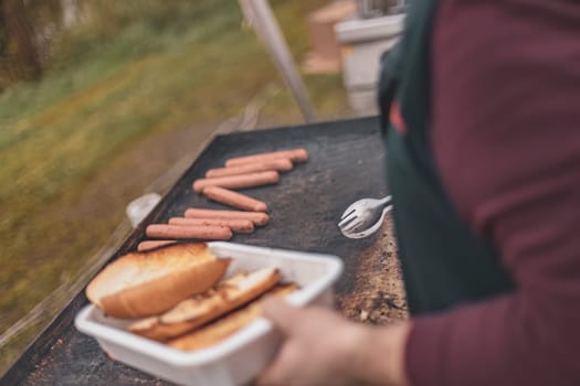 A person grilling hot dogs on a barbecue while holding a tray filled with sizzling sausages.