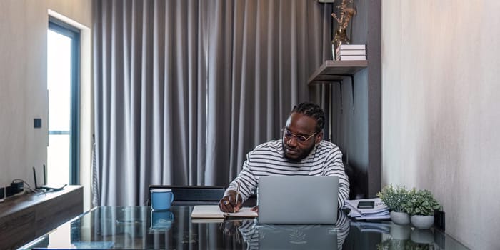 African American man working with laptop computer remote while sitting at glass table in living room. Black guy do freelance while taking notes work at home office.