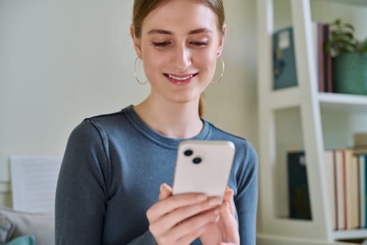 Happy smiling female teenager using smartphone, sitting on couch at home, girl 16,17, 18 years old texting reading messages. Modern digital technologies for communication, leisure, learning, shopping