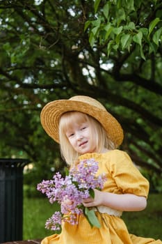 A little girl in a yellow dress and straw hat wearing a bouquet of lilacs. A walk in a spring park, blossoming lilacs