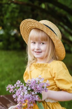 A little girl in a yellow dress and straw hat wearing a bouquet of lilacs. A walk in a spring park, blossoming lilacs