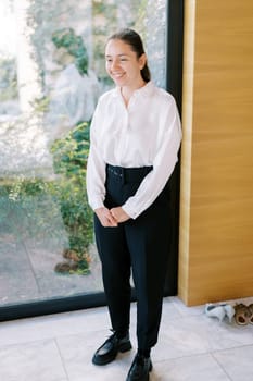 Smiling young woman stands near a panoramic window and looks away. High quality photo