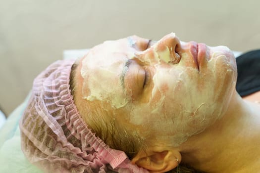 A man seated while a cosmetologist applies a facial mask to his face during a spa treatment session.