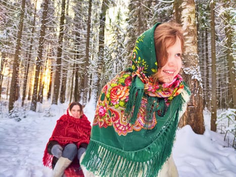 Mother and daughter in shawls are sledding in the winter forest. Ethnic clothes and fun on Shrovetide carnival Maslenitsa