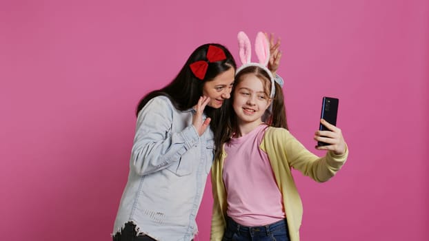 Playful cheery mother and girl smiling for photos on smartphone, having fun embracing each other in studio. Little child an her mom posing for pictures, fooling around. Camera B.