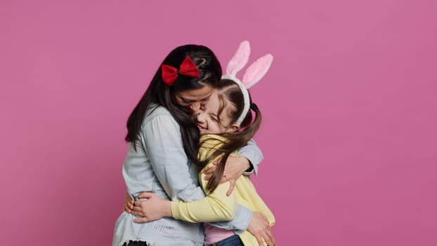 Lovely schoolgirl with bunny ears an her mom waving on camera, having fun and laughing against pink background. Cheerful mother and her daughter embracing and kissing each other. Camera A.