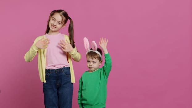 Joyful little kids waving in front of camera during easter holiday, smiling and wearing bunny ears. Brother and sister toddlers greeting someone in studio, adorable happy children. Camera A.