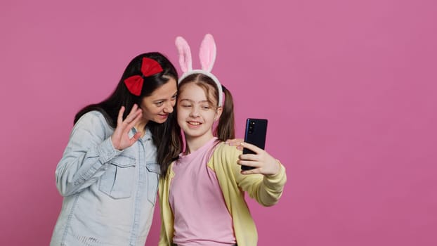 Playful cheery mother and girl smiling for photos on smartphone, having fun embracing each other in studio. Little child an her mom posing for pictures, fooling around. Camera A.