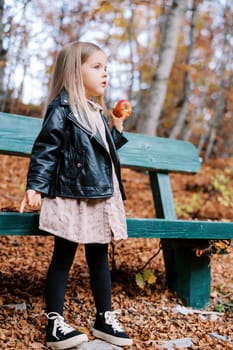 Little girl with an apple in her hand stands near a wooden bench in the autumn forest. High quality photo