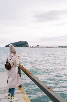 Young woman stands on the pier and takes pictures of the sea and mountains. Back view. High quality photo