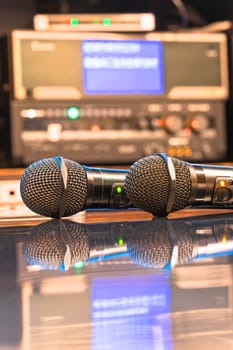 Two black microphones in a Japanese karaoke room reflecting on a table with a Karaoke machine and a controller adorned with LED-backlit buttons for adjusting microphone volume, echo and melody.