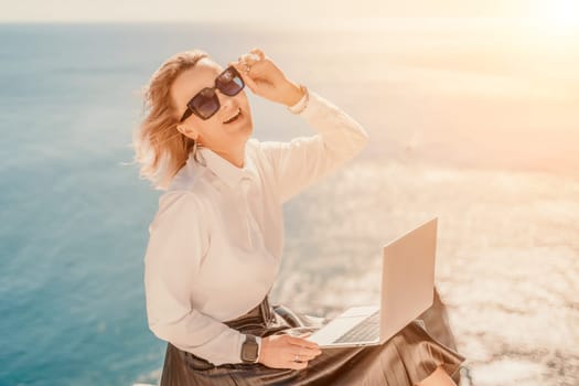 Business woman on nature in white shirt and black skirt. She works with an iPad in the open air with a beautiful view of the sea. The concept of remote work