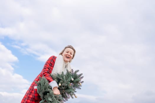Blond woman holding Christmas tree by the sea. Christmas portrait of a happy woman walking along the beach and holding a Christmas tree in her hands. Dressed in a red coat, white suit