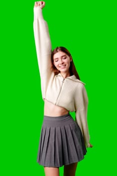 A cheerful young woman with long hair is captured mid-celebration, raising her arm high in a gesture of victory or excitement. She is dressed casually in a white sweater and a grey pleated skirt. The vivid green backdrop accentuates the positive and energetic mood conveyed by her stance.