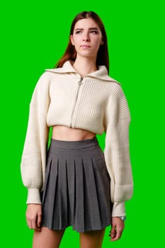 A young woman stands confidently against a vivid green background, dressed in a casual yet fashionable white cropped hoodie and a gray pleated skirt. Her expression is serene with a hint of contemplation, and her body language exudes a relaxed poise.