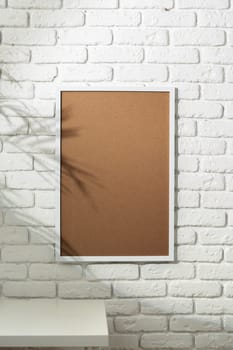 A clear shadow of palm leaves is cast upon a framed picture hanging on a white painted brick wall, creating an interesting mix of texture and patterns. A small potted plant next to a stack of books sits on a white surface in the foreground, adding a touch of greenery to the serene setting.