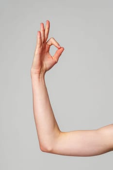 A close-up view of a persons arm extended upwards, with the hand forming an OK sign by connecting the thumb and index finger, against a simple, uncluttered grey backdrop.