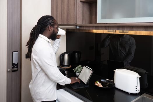 African American man working online on digital tablet and drinking coffee while at kitchen.