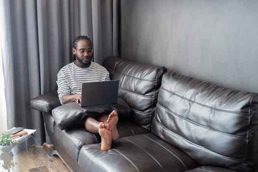 Young African American man holding laptop relaxing sitting on sofa working remote at home.