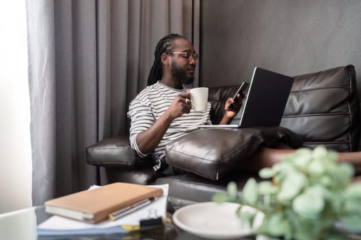 Young African American man holding laptop relaxing sitting on sofa working remote at home.