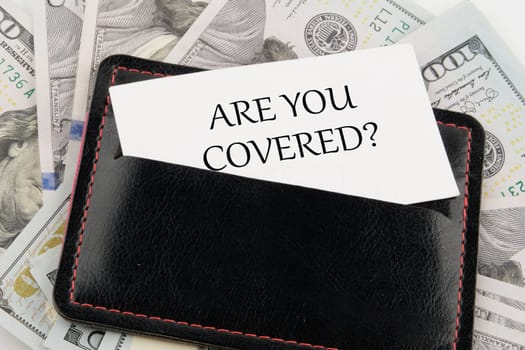 ARE YOU COVERED text written on a business card in a leather accessory on the background of banknotes
