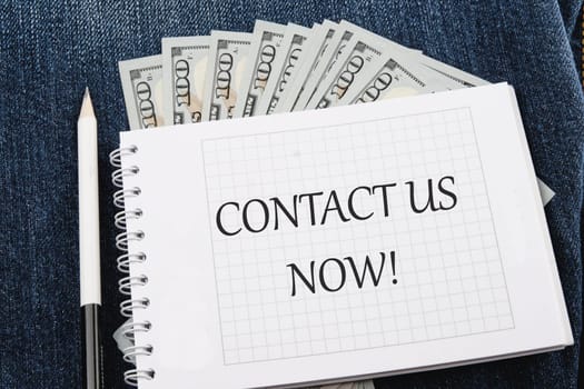 CONTACT US NOW word inscription on a notebook lying on jeans with dollar bills