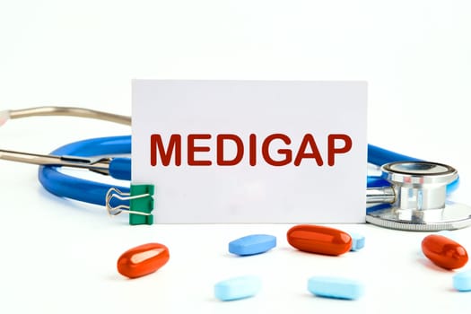 Medical concept. MEDIGAP on a white business card on a white background next to a stethoscope and pills, vitamins