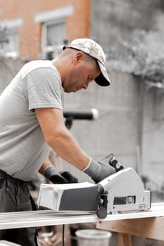 One young Caucasian recognizable man in a cap, uniform and gray gloves saws a board with an electric saw while standing in the backyard of a house on a summer day, close-up side view.
