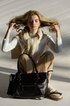 beautiful curly blond hair woman posing with a small shopper bag sitting on the floor. Model wearing stylish white sweater, classic trousers and loafer shoes