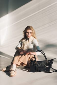 beautiful curly blond hair woman posing with a small shopper bag sitting on the floor. Model wearing stylish white sweater, classic trousers and loafer shoes