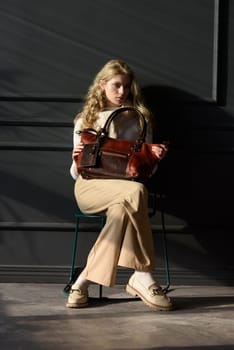 beautiful curly blond hair woman posing with a brown shopper bag near gray wall. Model wearing stylish white sweater, classic trousers and loafer shoes