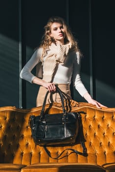 beautiful curly blond hair woman posing with a small black shopper bag near vintage volor sofa. Model wearing stylish white sweater and classic trousers