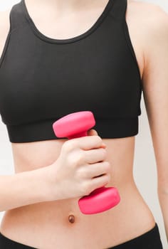 Young beautiful caucasian teen girl in a fitness suit holds one kilo pink dumbbell with one hand against the background of a white wall, close-up side view. Concept of sport, fitness and modern lifestyle.