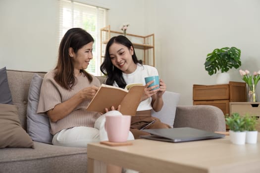 Happy senior mother with adult daughter sitting on couch and holding cups with coffee or tea at home. Enjoy family concept.