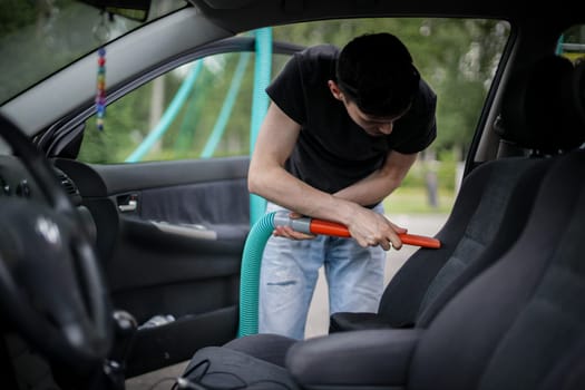 A handsome young caucasian brunette guy holds a tube with a nozzle in his hands and vacuums the seat in the car, side view close-up.