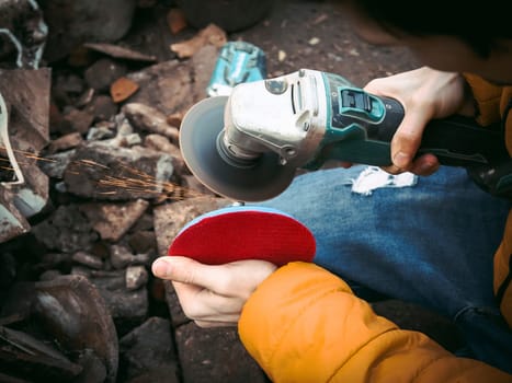 The hands of a young caucasian man in a yellow jacket are holding a drill and polishing with a disc in the backyard of a house with garbage on the ground, close-up side view. The concept of the polishing process.