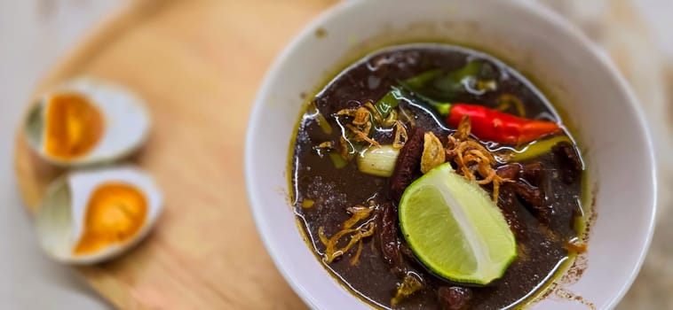 Rawon or Indonesian black beef soup, black color are from indonesian nut called kluwek. Served with lime, chili paste, salted egg, and tempe cracker. Perfect for recipe, article, or any cooking contents or blog