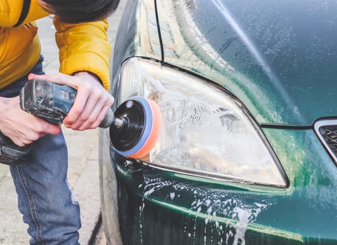 The hands of a young caucasian guy are drilling a drill with a disk with a sponge and polishing the headlight of his car with a detergent soap, close-up side view.The concept of a home car wash and cleaning with polishing car headlights.