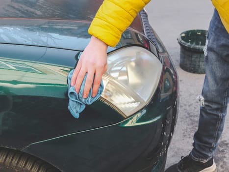A young caucasian guy in a yellow jacket holds blue rags in his hands and wipes the headlight of his car on a city street in front of the house, close-up side view. The concept of home car wash and cleaning with polishing car headlights.