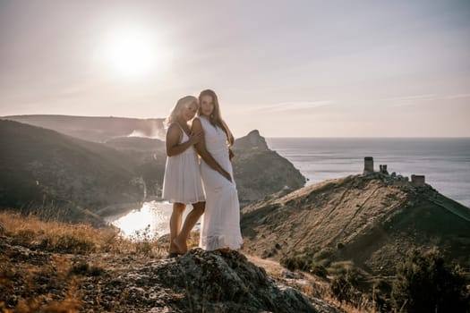 Two young girls are standing on a hillside, one of them wearing a white dress. The sun is shining brightly, creating a warm and inviting atmosphere. The girls seem to be enjoying their time together