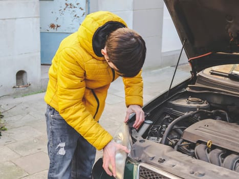 A young caucasian guy in a yellow jacket inspects an internal breakdown with an open car hood of his car on a city street in front of an old house, close-up side view. Engine concept for home repair.