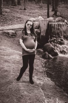 Monochrome photo of a pregnant girl in the park relaxing on a summer day by a waterfall wearing sunglasses