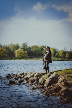 A pregnant girl in sunglasses stands on the shore of a lake against the background of a forest on a summer day