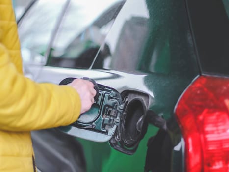 The hand of a young caucasian man in a yellow jacket holds the gas tank cap of a car at a city gas station, close-up side view. Gas station concept, service.