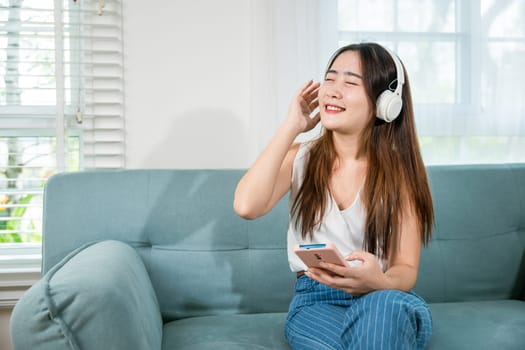 Beautiful young woman wear headphones listen music application on smartphone, Happy female wearing earphone listening to music with smart mobile phone and relaxing on sofa in living room at home