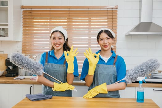 Two Asian young cleaning service women on kitchen counter with duster foggy spray and rag showcase efficient housework teamwork. Clean portrait two uniform maid working smiling employee.